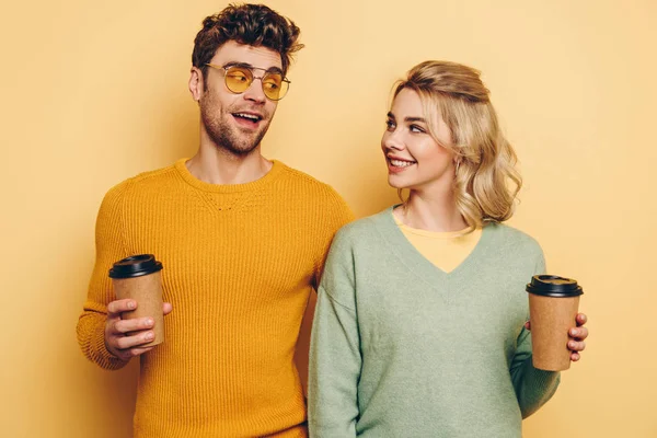 Happy man and woman looking at each other while holding coffee to go on yellow background — Stock Photo