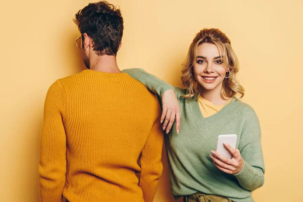 Vue arrière de l'homme debout près d'une fille souriante en utilisant un smartphone sur fond jaune — Photo de stock