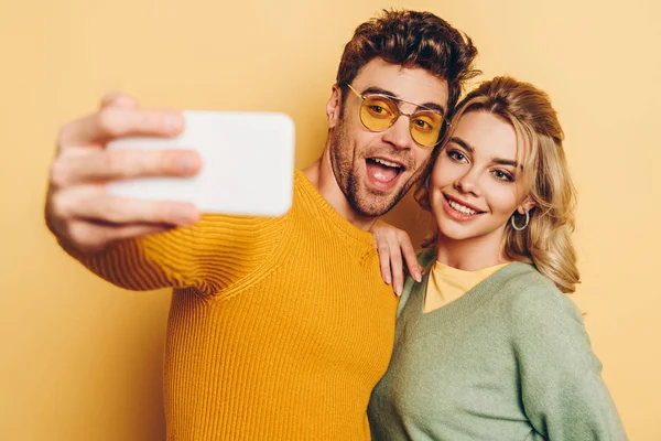 Hombre excitado tomando selfie en el teléfono inteligente con novia atractiva en el fondo amarillo - foto de stock