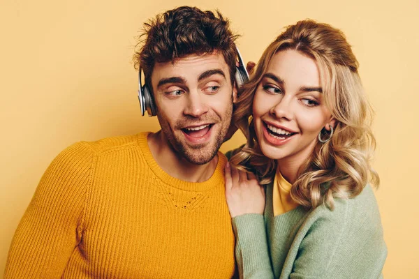 Menina feliz abraçando namorado enquanto ouve música em fones de ouvido sem fio juntos no fundo amarelo — Fotografia de Stock