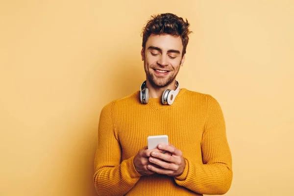 Uomo sorridente con cuffie wireless sul collo chatta su smartphone su sfondo giallo — Foto stock