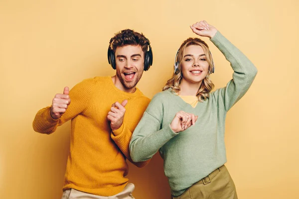 Couple heureux dansant tout en écoutant de la musique dans des écouteurs sans fil sur fond jaune — Photo de stock