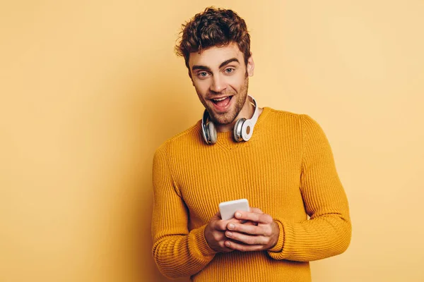 Hombre guapo con auriculares inalámbricos en el cuello usando teléfono inteligente y sonriendo a la cámara en el fondo amarillo - foto de stock