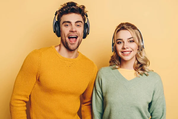 Felice uomo e donna che sorridono alla fotocamera mentre ascoltano musica in cuffie senza fili su sfondo giallo — Foto stock