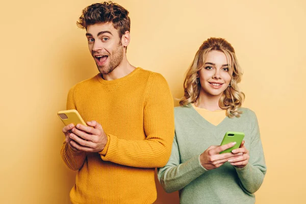 Alegre hombre y mujer sonriendo a la cámara mientras que los mensajes en los teléfonos inteligentes en el fondo amarillo - foto de stock