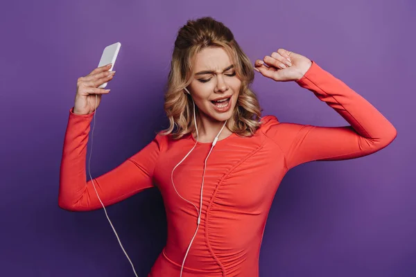 Весела дівчина тримає смартфон і танцює під час прослуховування музики в навушниках на фіолетовому фоні — стокове фото