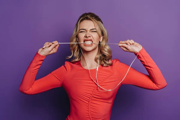 Emocionado chica mordiendo alambre de auriculares mientras escucha música sobre fondo púrpura - foto de stock