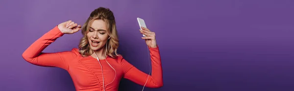 Plan panoramique de fille excitée dansant tout en écoutant de la musique dans les écouteurs et en tenant smartphone sur fond violet — Photo de stock