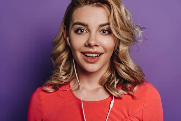 Cheerful girl listening music in earphones while smiling at camera on purple background — Stock Photo