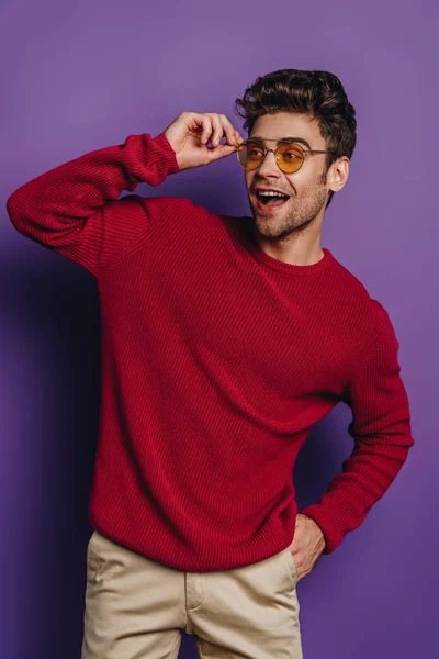 Hombre alegre tocando gafas mientras está de pie con la mano en el bolsillo y mirando hacia otro lado sobre fondo púrpura - foto de stock