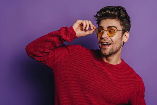 Uomo allegro toccare gli occhiali mentre guardando lontano su sfondo viola — Foto stock