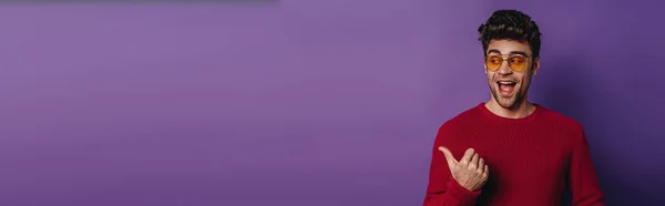 Панорамный снимок веселого молодого человека, указывающего пальцем и отводящего взгляд на фиолетовый фон — стоковое фото