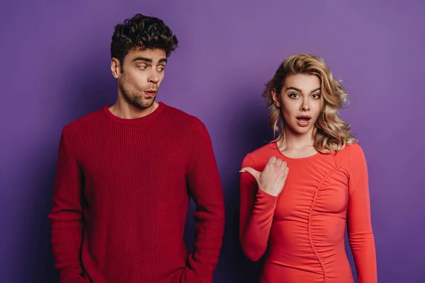 Shocked girl pointing with thumb at discouraged boyfriend on purple background — Stock Photo