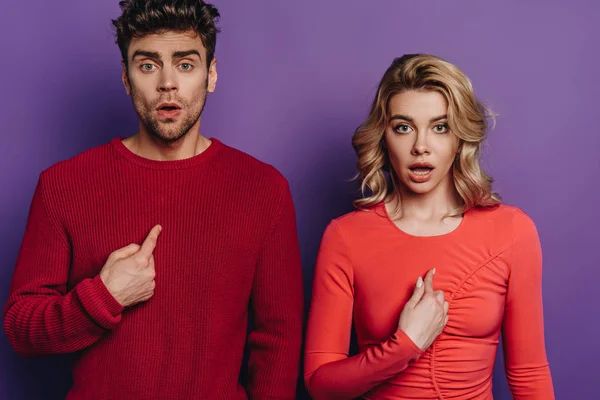 Surprised man and woman pointing with fingers at themselves while looking at camera on purple background — Stock Photo