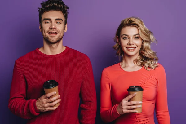 Sorrindo casal olhando para a câmera enquanto segurando café para ir no fundo roxo — Fotografia de Stock