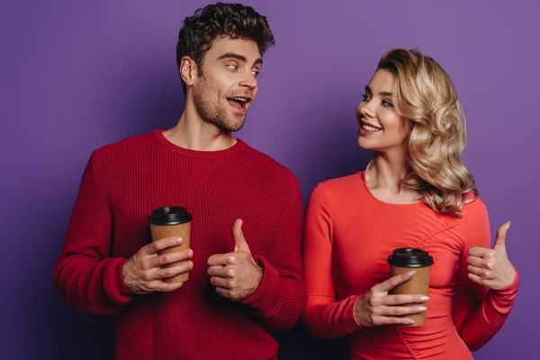 Happy couple showing thumbs up while holding coffee to go on purple background — Stock Photo