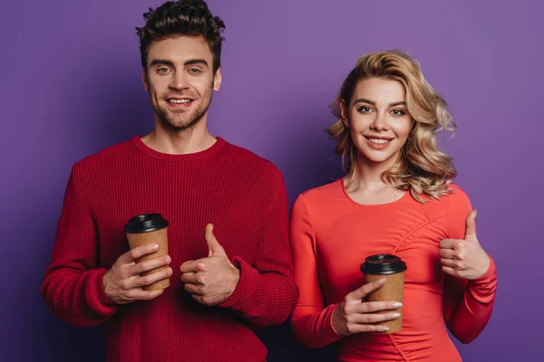 Cheerful couple showing thumbs up while holding coffee to go on purple background — Stock Photo