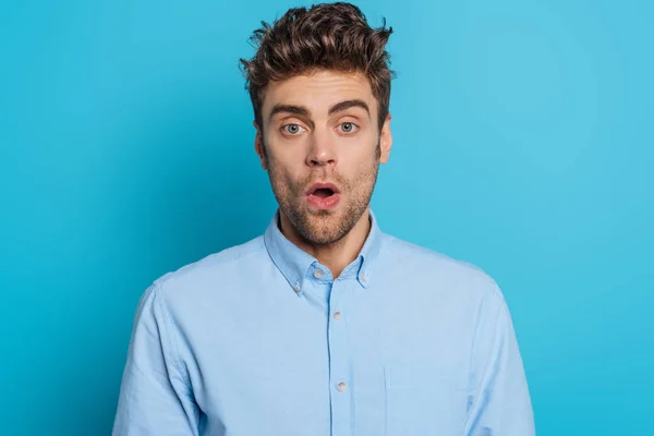 Choqué jeune homme regardant la caméra tout en se tenant debout avec la bouche ouverte sur fond bleu — Photo de stock