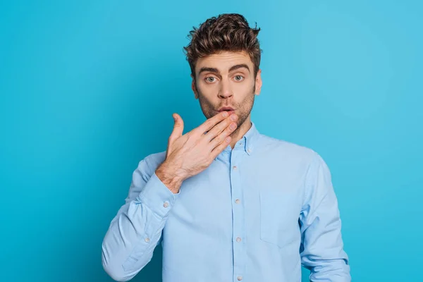 Überraschter Mann, der Mund mit der Hand bedeckt, während er in die Kamera auf blauem Hintergrund schaut — Stockfoto