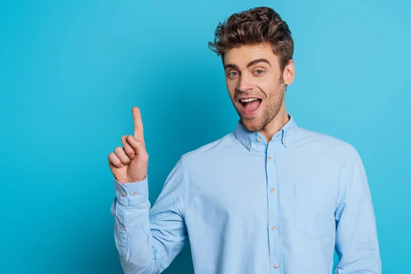 Cheerful man showing idea gesture while smiling at camera on blue background — Stock Photo