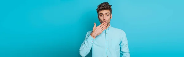 Panoramic shot of shocked man covering mouth with hand on blue background — Stock Photo