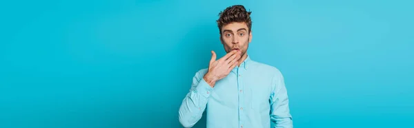 Plano panorámico del hombre sorprendido cubriendo la boca con la mano sobre fondo azul - foto de stock