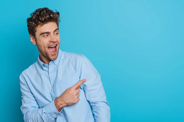 Excited young man looking away and laughing while pointing with finger on blue background — Stock Photo