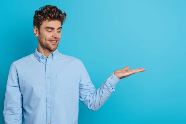 Sorridente jovem olhando para o braço aberto no fundo azul — Fotografia de Stock