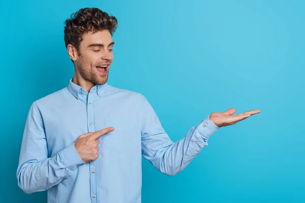 Fröhlicher junger Mann zeigt mit dem Finger auf offenen Arm auf blauem Hintergrund — Stockfoto