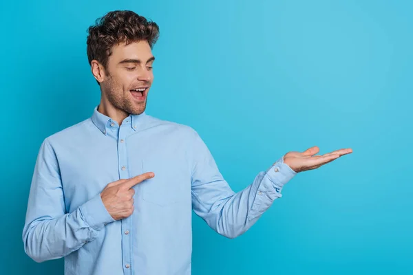 Alegre joven apuntando con el dedo en el brazo abierto sobre fondo azul - foto de stock