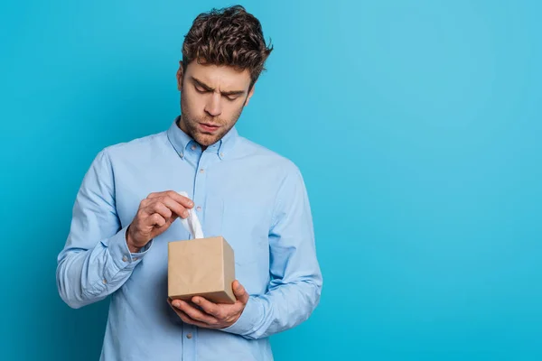 Aufgebrachter junger Mann nimmt Papierserviette aus Packung auf blauem Hintergrund — Stockfoto