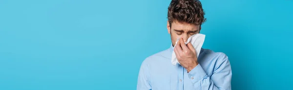 Panoramic shot of diseased man sneezing in paper napkin on blue background — Stock Photo