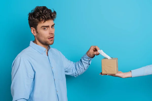 Abgeschnittene Ansicht einer Frau, die einem verärgerten Mann eine Packung Papierservietten gibt und dabei in die Kamera auf blauem Hintergrund schaut — Stockfoto