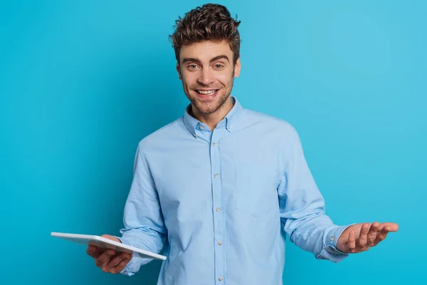 Веселый молодой человек, стоящий с открытой рукой, держа в руках цифровой планшет и улыбаясь в камеру на синем фоне — стоковое фото