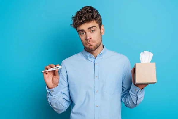 Diseased young man holding pack of paper napkins and showing thermometer on blue background — Stock Photo