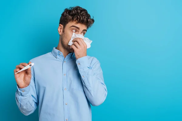 Malade jeune homme essuyant nez avec serviette en papier et montrant thermomètre sur fond bleu — Photo de stock