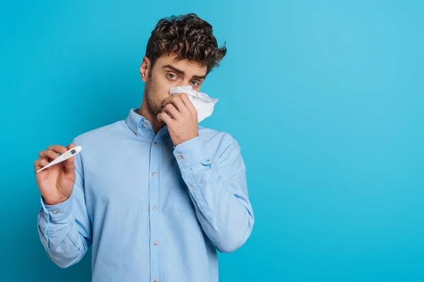 Malade jeune homme essuyant nez avec serviette en papier et montrant thermomètre sur fond bleu — Photo de stock