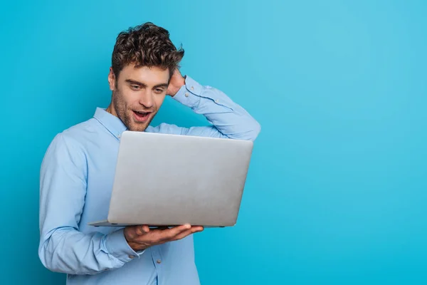 Überraschter junger Mann berührt Kopf, während er auf Laptop auf blauem Hintergrund schaut — Stockfoto