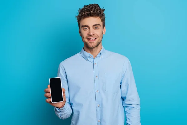 Joven guapo sonriendo a la cámara mientras muestra el teléfono inteligente con pantalla en blanco sobre fondo azul - foto de stock