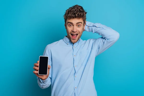 Sorprendido joven tocando la cabeza mientras muestra el teléfono inteligente con pantalla en blanco sobre fondo azul — Stock Photo