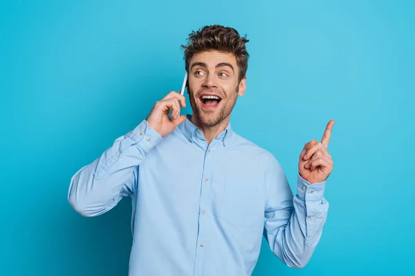 Joven emocionado mostrando gesto de idea y mirando hacia otro lado mientras habla en el teléfono inteligente sobre fondo azul — Stock Photo