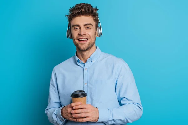 Joven alegre sonriendo a la cámara mientras escucha música en auriculares inalámbricos y sosteniendo el café para ir sobre fondo azul - foto de stock