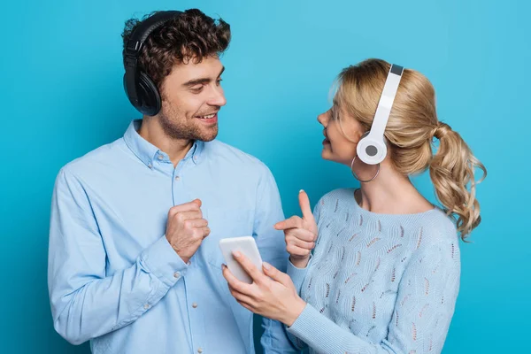 Pareja positiva en los auriculares mirándose entre sí mientras que la chica señala con el dedo en el teléfono inteligente sobre fondo azul - foto de stock