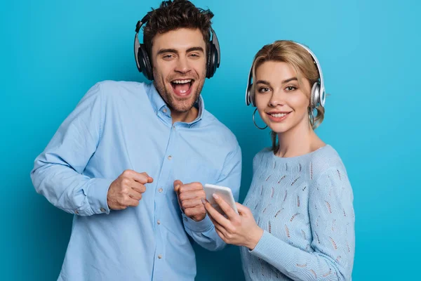 Alegre hombre y mujer mirando a la cámara mientras escucha música en auriculares inalámbricos sobre fondo azul - foto de stock