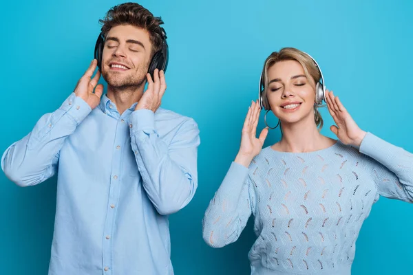 Happy man and woman listening music in wireless headphones with closed eyes on blue background — Stock Photo