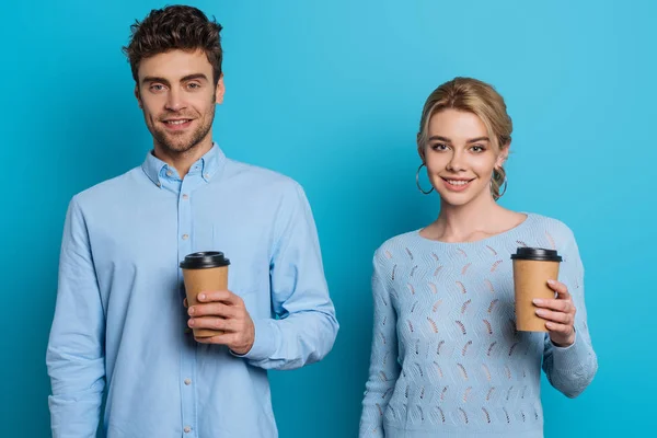 Homem positivo e mulher segurando copos descartáveis enquanto sorrindo para a câmera no fundo azul — Fotografia de Stock