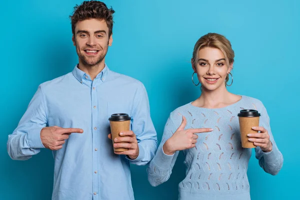 Sonriente hombre y mujer señalando con los dedos a las tazas desechables mientras mira a la cámara en el fondo azul - foto de stock