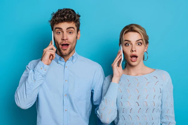 Surpris homme et femme regardant la caméra tout en parlant sur les smartphones sur fond bleu — Photo de stock