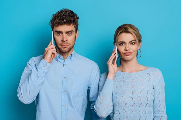 Skeptical man and woman looking at camera while talking on smartphones on blue background — Stock Photo