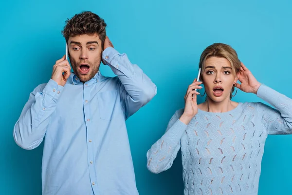 Shocked man and woman touching heads while talking on smartphones on blue background — Stock Photo